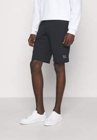 Pantaloni corti blu navy con una texture liscia, vita elastica e logo EA7 ricamato. Abbinati a sneakers bianche e una maglietta bianca a maniche lunghe.
