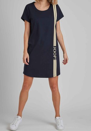Kvinde iført en marineblå kortærmet kjole med lodrette beige striber og tekst, kombineret med hvide sneakers, stående mod en ensfarvet baggrund.