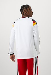 Ung man med flätat hår, klädd i en vit långärmad tröja med svarta, röda och gula axelränder samt röda träningsbyxor med vita och svarta ränder.