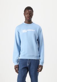Šviesiai mėlynas megztinis su guma apyrankėmis ir apatine dalimi, ant krūtinės raudonai išsiuvinėtas balta „KARL LAGERFELD JEANS“ tekstas. Dėvima su tamsiai džinsais.