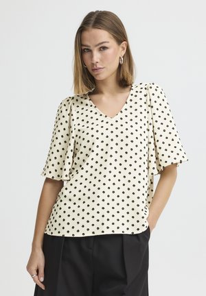 Frau mit schulterlangem Haar, die eine cremefarbene Bluse mit schwarzen Punkten und Kragenärmeln trägt, kombiniert mit schwarzen Hosen, steht mit einer Hand in der Hosentasche.