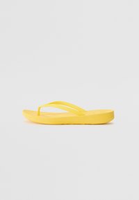 Keltainen flip-flop-sandaali, jossa on litteä pohja, kaksi ohutta hihnaa, sileä pinta ja yksinkertainen muotoilu. Sopii rentoon käyttöön.