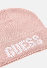 Rožinė megzta skrybėlė su sulankstytu kraštu, kuriame priekyje puikuojasi balta "GUESS" užrašas, papuoštas sidabriniais kaiščiais. Minkšta tekstūra, tvirtas prigludimas.