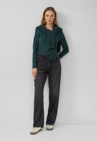 Dunkelgrünes, geripptes Langarmoberteil mit einem gestrickten Schal, kombiniert mit hoch taillierten schwarzen Jeans und hellen Sneakers.