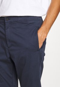 Homme portant un pantalon bleu marine avec la main dans la poche droite, associé à une chemise blanche rentrée.