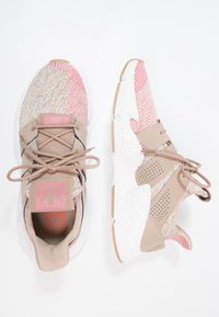 Beżowe i różowe buty sportowe z teksturowaną cholewką, białą podeszwą oraz połączeniem materiałów dzianinowych i syntetycznych. Zawierają tonowane sznurowadła oraz warstwowy design.