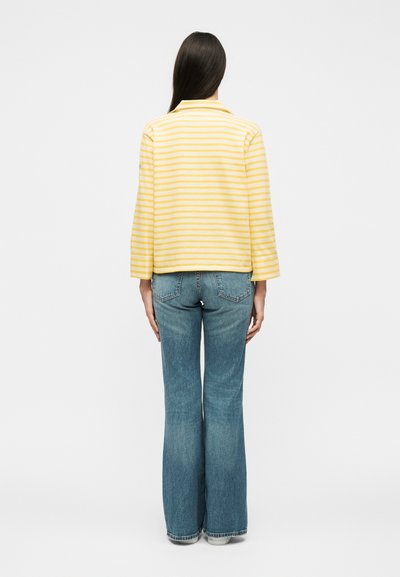 Γυναίκα με μακριά σκούρα μαλλιά, φοράει κίτρινη και λευκή ριγέ μπλούζα με μακριά μανίκια και μπλε τζιν με φαρδιά μπατζάκια, στέκεται με την πλάτη προς τα εμπρός.