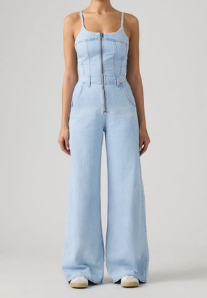 Kvinde iført lyseblåt denim jumpsuit med lynlås foran, tynde stropper, vide ben og hvide sneakers står mod en ensartet baggrund.