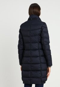 Mörkblå vadderad jacka med hög krage, med en kviltad design, långa ärmar och knappstängning vid halsen.