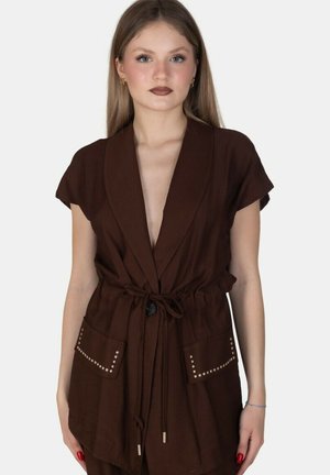 Giovane donna con lunghi capelli biondi indossa una giacca marrone a maniche corte con cintura, tasche con borchie e smalto rosso sulle unghie su sfondo bianco.