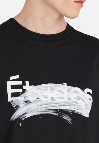 T-shirt en coton noir avec un design graphique blanc audacieux, portant le texte "Études" partiellement obscurci par un coup de pinceau.