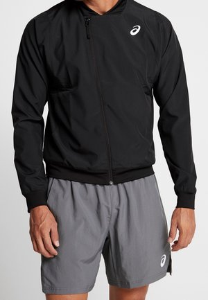 Homme portant une veste de sport noire zippée et un short de sport gris avec des logos blancs sur la poitrine et la jambe sur fond blanc.
