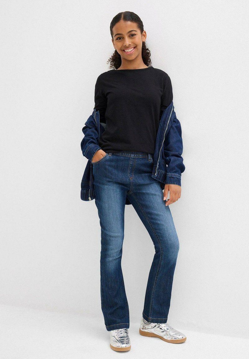 Jeans in denim scuro con una vestibilità dritta e una vita elastica, abbinati a un top nero a maniche lunghe e a un giubbotto di denim blu.