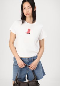 Valkoinen puuvillapaitapusero, jossa on punainen cowboyn saappaan grafiikka edessä. Yhdistettyinä kuluneisiin farkkushortseihin ja tummaan käsilaukkuun.