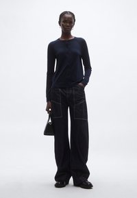 Tmavomodrý kardigán s dlhými rukávmi a gombíkmi, kombinovaný so širokými tmavými džínami s prednými vreckami a kontrastným prešitím. Drží malú čiernu tašku.