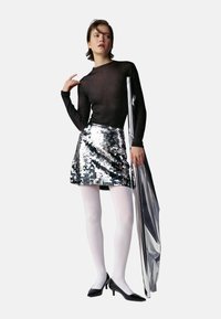 Haut noir à manches longues, mini-jupe argentée à paillettes, collants blancs et talons noirs. Mannequin tenant un accessoire métallique argenté, fond simple.