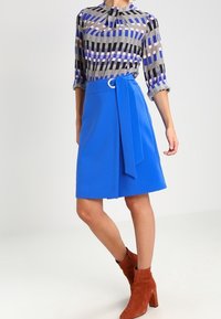 Kvinna i en blå omslagskjol med knyt-detalj, mönstrad långärmad blus och bruna mocka ankelstövlar, som står mot en enfärgad bakgrund.