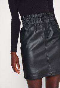 Černá kožená sukně s řaseným pasem, má dvě boční kapsy a dva přední knoflíky, hladká textura a rovný střih.