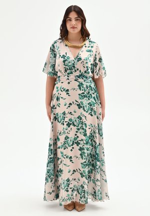Femme portant une robe maxi beige avec imprimé floral vert, manches courtes volantées, encolure en V, collier doré et chaussures pointues marron.