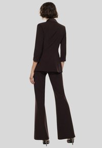 Completo marrone su misura con giacca aderente e pantaloni svasati. La giacca ha colletto a revers e maniche a tre quarti. Tessuto dalla superficie liscia.