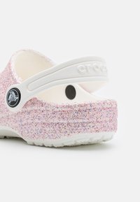 Glitterige roze crocs met een witte band en een cirkelvormig logo-accent, voorzien van een gestructureerde zool en ronde vorm. Glad en lichtgewicht ontwerp.
