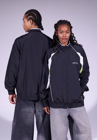 Jaqueta corta-vento preta com detalhes em branco e verde-lima, punhos elásticos e ajuste relaxado. Apresenta colarinho alto e fecho com zíper.