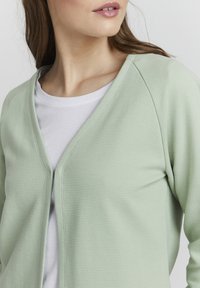 Světl zelený kordový cardigan s výstřihem do V a volným předním otvorem, překrytý bílým tričkem. Měkká textura s volnočasovým designem.