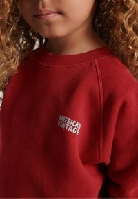 Raudona džemperis, pagamintas iš minkštos medžiagos, su apvalia iškirpte. Ant krūtinės yra baltas "AMERICAN VINTAGE" logotipas ir matomos siuvimo detalės.