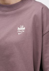 Vijoličast pulover s belim besedilom "Café Swoosh Fresh Daily" nad logotipom Nike swoosh na prsih.