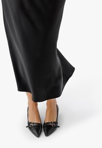 Mujer que lleva tacones negros de punta con correas de doble hebilla y una larga falda negra fluida, de pie sobre un suelo blanco.