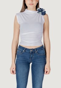 Top bianco senza maniche con tessuto arricciato, collo alto e un accento floreale blu sulla spalla sinistra, abbinato a jeans blu aderenti.