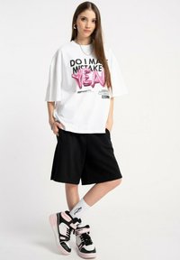 Młoda kobieta z długimi włosami, ubrana w białą, oversize'ową koszulkę z napisem "CZY POPEŁNIAM BŁĘDY YEA", czarne szorty, białe skarpetki i czarno-różowe trampki.
