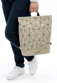 Henkilö, joka käyttää tummia farkkuja ja valkoisia tennareita, pitää kädessään beigeä, geometrisesti kuvioitua Suri Frey -laukkuja sen kahvasta.