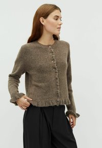 Barna gyapjú kardigán kerek nyakkivágással, gombos elülső résszel és bolyhos szegéllyel. Hosszú ujjakkal rendelkezik, bolyhos mandzsettákkal.