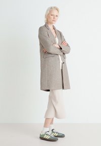 Sivý štruktúrovaný dlhý kabát cez krémový top, skombinovaný s béžovou sukňou a zelenými teniskami. Modelka stojí s prekríženými rukami pred svetlým pozadím.