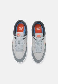 Ténis Nike SB em camurça cinza-claro com detalhes em cinza-escuro. Apresenta logótipos laranja, forro azul e atacadores cinza planos. Sola branca.