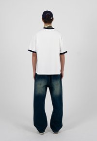 Homme portant une chemise blanche à manches courtes avec un liseré noir, un jean large foncé, une casquette de baseball bleue marine et une écharpe à motifs, debout de dos.