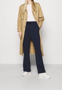 Beige trench coat, lightweight fabric, double-breasted, paired with navy wide-leg pants, elastic waistband, white sneakers, smooth texture.