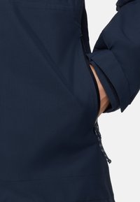 Mano nella tasca di un giubbotto blu scuro con zip visibile e polsini regolabili, su sfondo bianco.