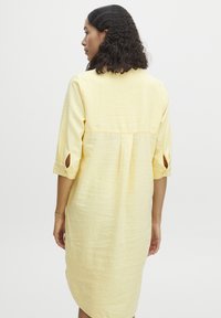 Robe jaune, midi, à coupe décontractée, avec des manches trois-quarts et des poignets boutonnés, fabriquée en tissu léger et texturé.