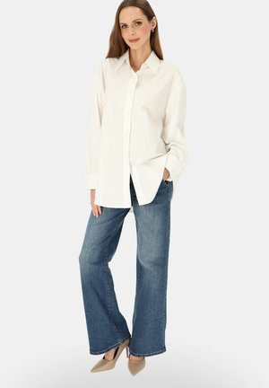 Kobieta stojąca, ubrana w białą, oversize koszulę na guziki, szerokie niebieskie dżinsy i beżowe szpilki, z jedną ręką w kieszeni.