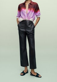 Persona che indossa una camicia ombre viola-rossa parzialmente sbottonata, pantaloni neri in pelle a vita alta e sandali neri con cinturino intrecciato.