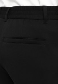 Zwarte broek met een gestructureerde stof, een gladde tailleband en een enkele achterzak met een subtiele stiknaad.