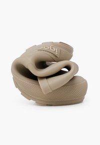 Chaussure en caoutchouc beige avec un design torsadé, surface texturée et accents surélevés. Présente un logo de marque sur le côté et une forme cintrée.