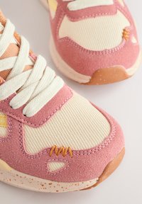 Les baskets présentent une tige en suède rose avec des accents en textile crème, des lacets blancs et des coutures orange. Semelle en caoutchouc texturée avec des détails tachetés.