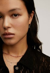 Pendientes de plata con un diseño circular, collares en capas—uno con cuentas rosas y otro con un pequeño charm en forma de "L"—sobre una blusa de encaje negra.