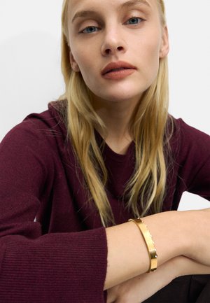 Złota metalowa bransoletka na nadgarstku, o gładkim wykończeniu i małych okrągłych akcentach. Modelka nosi bordowy sweter dziany.