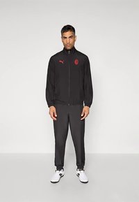 Black sports jacket with red Puma logo, zip front, elastic cuffs, paired with matching black pants and white athletic shoes.