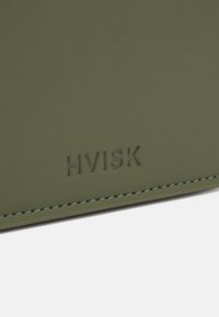 Portfel z oliwkowej sztucznej skóry z wytłoczonym logo "HVISK", o delikatnej fakturze i dopasowanym przeszyciu wzdłuż krawędzi.
