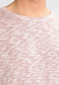 Ljusrosa stickad tröja med en strukturerad, fläckig mönstring, rund halsringning och mjukt tyg. Närbild på texturen och halsringningen.